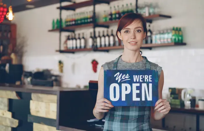 Female cafe owner smiling and holding a blue “Yes We’re Open” sign inside cozy coffee shop with bottles on shelves